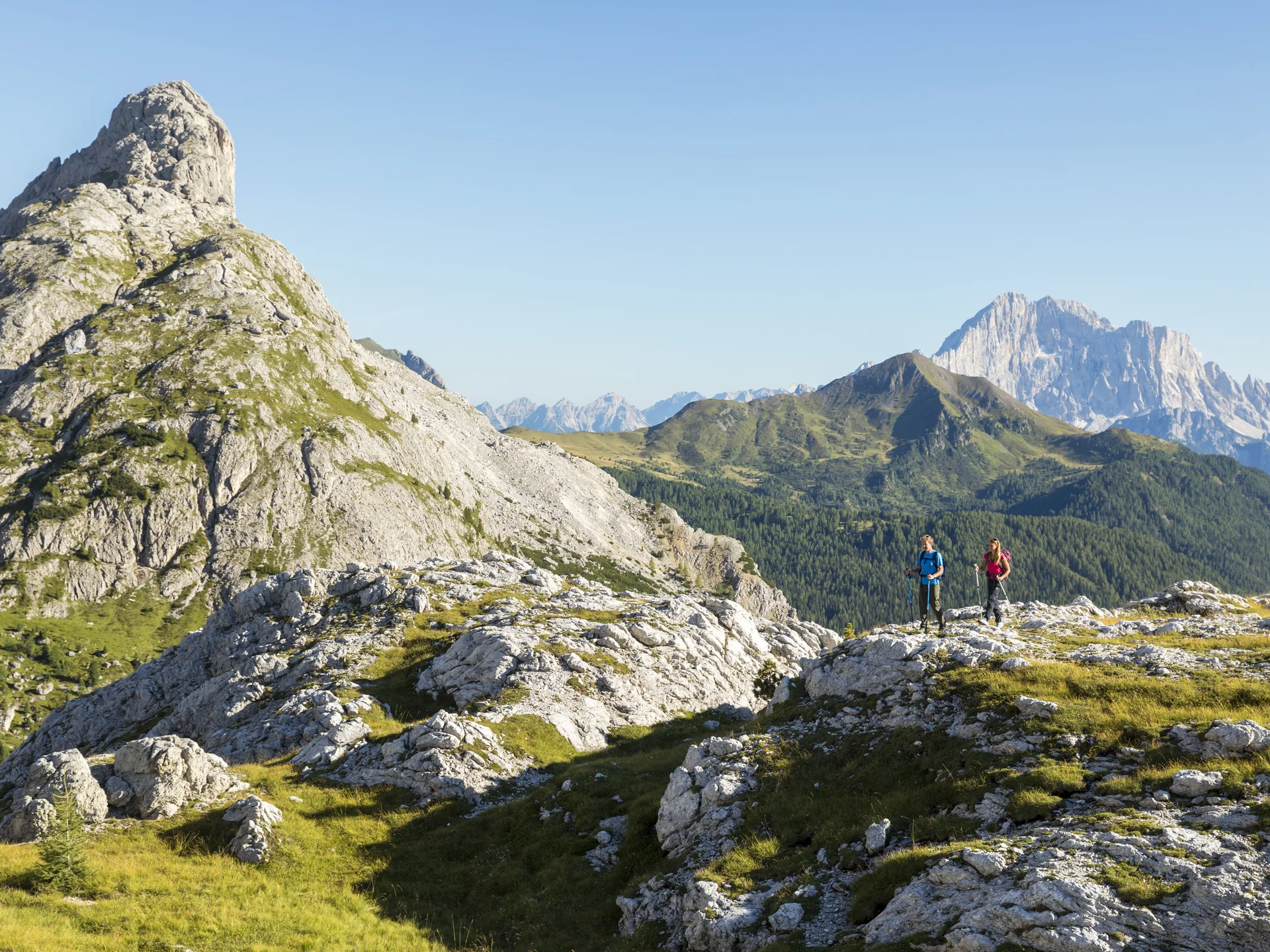 Il tuo Chalet ad Arabba: Chalet Barbara, charme alpino. Due escursionisti su un sentiero roccioso tra montagne verdi e cielo sereno