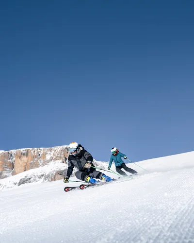 Il tuo Chalet ad Arabba: Chalet Barbara, charme alpino. Due sciatori che scendono una pista innevata con montagne sullo sfondo